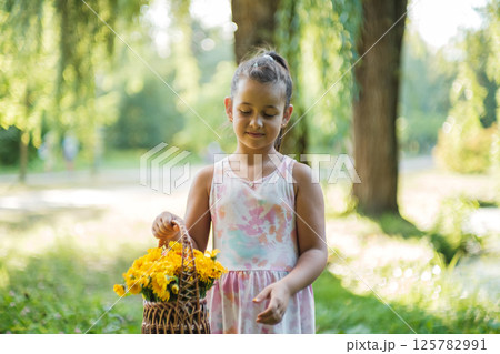 In a lovely park, a girl in a dress carries a basket of yellow flowers. 125782991