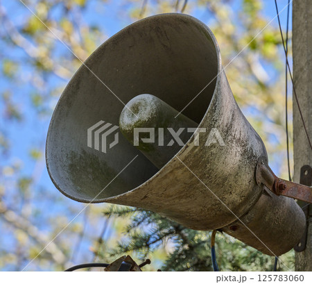 An old, dirty, grey loudspeaker is mounted on a pole. In the background, there is a blurred image of the sky and tree branches. An old, dirty, grey loudspeaker is mounted on a pole. In the background, there is a blurred image of the sky and tree branches. 125783060