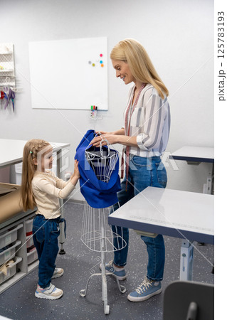 Blonde long-haired female dress designer in her studio with her daughter Blonde long-haired female dress designer in her studio with her daughter 125783193