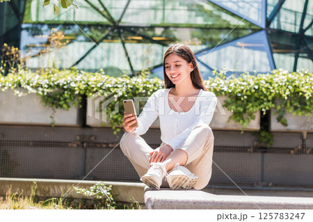 Relaxed young woman sitting outside with her phone, soaking in sunshine while managing wellness apps, chats, and peaceful productivity on the go Relaxed young woman sitting outside with her phone, soaking in sunshine while managing wellness apps, chats, and peaceful productivity on the go 125783247
