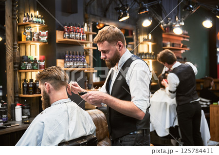 Professional barber using comb and electric shaver to cut side hair. Male customer getting stylish haircut. Side view of man on background of other hairdresser. Professional barber using comb and electric shaver to cut side hair. Male customer getting stylish haircut. Side view of man on background of other hairdresser. 125784411