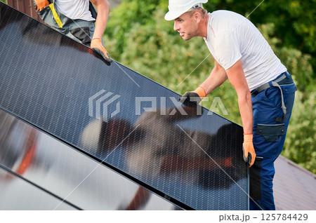 Workers building solar panel system on roof of house. Two men installers in helmets carrying photovoltaic solar module outdoors. Alternative, green and renewable energy generation concept. Workers building solar panel system on roof of house. Two men installers in helmets carrying photovoltaic solar module outdoors. Alternative, green and renewable energy generation concept. 125784429