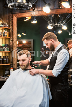 Professional barber shaving client's nape. Bearded handsome client getting stylish haircut. Confident man on regular appointment in barbershop. 125784444