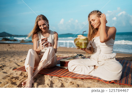 Two Young Women Relaxing at the Beach with Drinks and Smartphones 125784467
