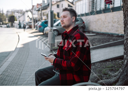 Male student with smartphone wearing plaid shirt sitting on bench while waiting for transport at tram or bus station.  125784797