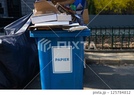 Blue trash bin labeled "PAPIER" for paper waste, overflowing with cardboard and paper 125784812