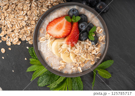 Cooked spelt in bowl. Oatmeal milk porridge with fruits and berries on dark background Cooked spelt in bowl. Oatmeal milk porridge with fruits and berries on dark background 125785717