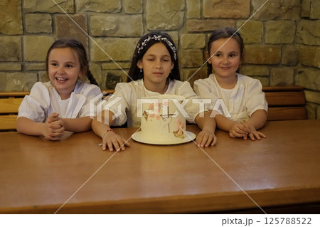 The cake decorates the center of the table, surrounded by joyful girls. The older sister and the twins admire each element of the dessert with fascination. 125788522