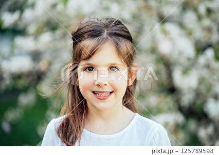 Cheerful Childhood Moment Among Blooming Trees. Smiling young girl with brown hair in white shirt standing outdoors in front of blooming spring trees 125789598