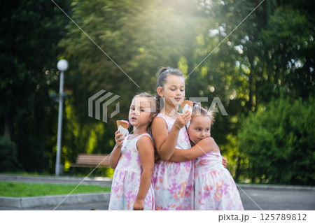 Three sisters, dressed in identical dresses, stand holding hands and enjoying ice cream.  125789812