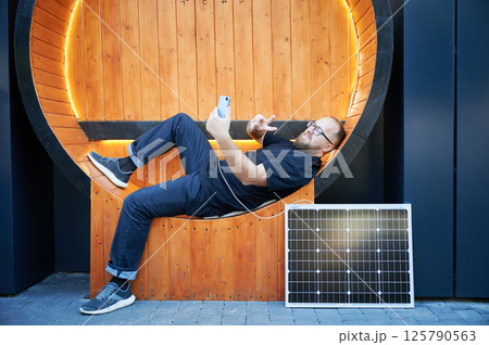 Bearded man using smartphone connected to photovoltaic solar panel. Integration of sustainable renewable energy into everyday life, demonstrating practical use of solar power for charging devices. Bearded man using smartphone connected to photovoltaic solar panel. Integration of sustainable renewable energy into everyday life, demonstrating practical use of solar power for charging devices. 125790563