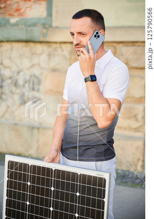 Handsome man using smartphone connected to photovoltaic solar panel. Integration of sustainable renewable energy into everyday life, demonstrating practical use of solar power for charging devices. Handsome man using smartphone connected to photovoltaic solar panel. Integration of sustainable renewable energy into everyday life, demonstrating practical use of solar power for charging devices. 125790566