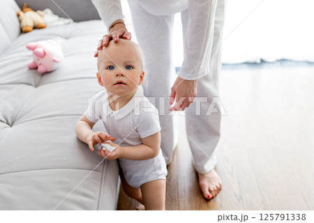 Toddler baby boy who learning to walk standing by couch with Woman. Baby development and parent support In bright white room at home Toddler baby boy who learning to walk standing by couch with Woman. Baby development and parent support In bright white room at home 125791338