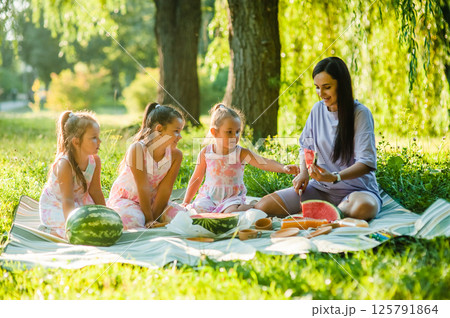 Cute little girls of 4-5 years old and their mother are eating a delicious watermelon against the background of green nature. Healthy lifestyle. Cute little girls of 4-5 years old and their mother are eating a delicious watermelon against the background of green nature. Healthy lifestyle. 125791864