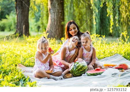 The girls and their mother are eating watermelon, their dresses are a little dirty. The twins do not pay attention, laughing together. The girls and their mother are eating watermelon, their dresses are a little dirty. The twins do not pay attention, laughing together. 125793152