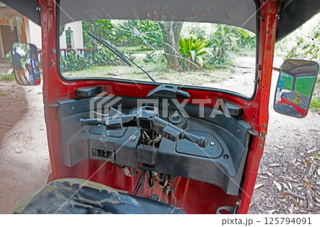 Steering controls and dashboard of a red tuk tuk in a tropical village with no people visible Steering controls and dashboard of a red tuk tuk in a tropical village with no people visible 125794091
