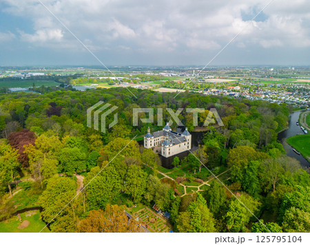 Aerial view of a historic castle surrounded by a forest, with vast green fields, winding waterways, and a river stretching across the flat Dutch countryside under a partly cloudy sky. 125795104