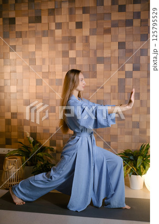 Yoga instructor in blue kimano at fitness gym . The girl does qigong yoga. The exercises are based on the movement of internal energy, the system of channels, energy centers, chakras Yoga instructor in blue kimano at fitness gym . The girl does qigong yoga. The exercises are based on the movement of internal energy, the system of channels, energy centers, chakras 125795529