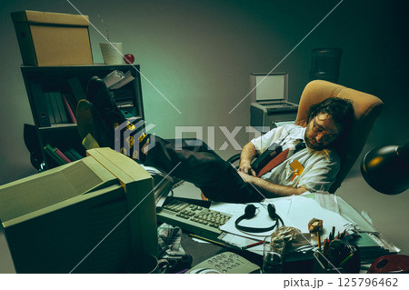 Man with glasses feel asleep in office chair with his legs on desk, surrounded by papers and computer. Overwhelming work and the need for rest. 125796462