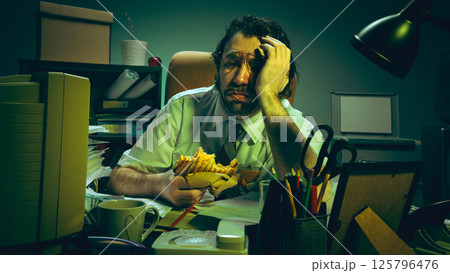 Tired employee holds sandwich with blank stare at chaotic desk surrounded by paperwork, showing burnout, exhaustion, stress emotions, pressure. 125796476