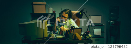 Man buried in documents, balances two phones while working under lamp in cluttered office, expressing burnout, fatigue, pressure. Man buried in documents, balances two phones while working under lamp in cluttered office, expressing burnout, fatigue, pressure. 125796482