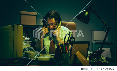 Stressed office worker holding two phones at messy desk in dimly lit, green-tinted room, communication overload, pressure, chaos of demanding job Stressed office worker holding two phones at messy desk in dimly lit, green-tinted room, communication overload, pressure, chaos of demanding job 125796495