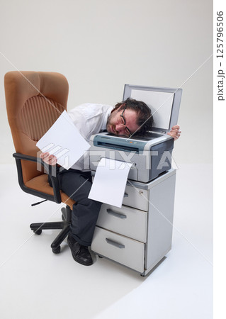 Man in shirt and tie lies over printer, holding paper, showing professional burnout, fatigue and frustration. Man in shirt and tie lies over printer, holding paper, showing professional burnout, fatigue and frustration. 125796506