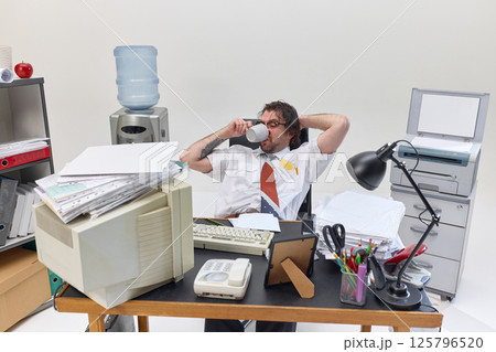 Office worker drinks coffee, reclining at cluttered desk surrounded by paperwork, showing fatigue, break and attempt to refocus during long day. 125796520