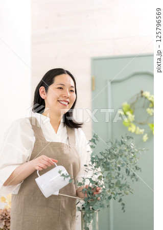 室内で植物の水やりをする笑顔の大人女性｜花屋園芸店教室イメージ｜ウェルビー女子｜ウェルビーイング 125796569