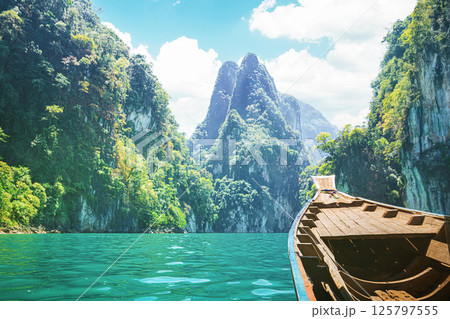 Travelers enjoy a cruise on a traditional longtail boat in Cheow Lan Lake, surrounded by towering limestone cliffs and lush greenery. The vibrant blue waters and scenic mountains 125797555
