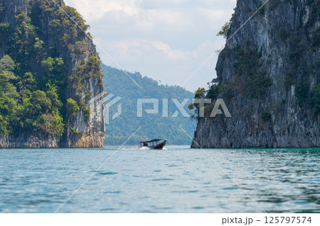 Small boat glides across the turquoise waters of Cheow Lan Lake, by towering limestone cliffs covered in lush greenery. Bright sunlight reflects on the lake's surface, enhancing the beauty of Khao Sok 125797574
