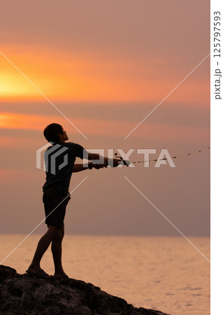 fisherman casting a spinning rod into the sea from a cliff against the background of an orange sea sunset, rear view close-up fisherman casting a spinning rod into the sea from a cliff against the background of an orange sea sunset, rear view close-up 125797593