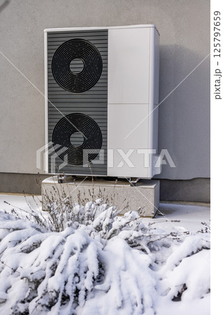 Close-up of a dual-fan outdoor heat pump on a gray wall with snow-covered bushes, highlighting modern winter energy efficiency. Close-up of a dual-fan outdoor heat pump on a gray wall with snow-covered bushes, highlighting modern winter energy efficiency. 125797659