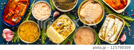 Enjoy an assortment of canned fish including salmon, mackerel, sprat, sardine, tuna, herring, and fish pate, displayed in open tins with spices and herbs on a blue background, banner 125798309