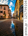 Cobblesone street among buildings under blue sky with white clouds in old town of Alba, Italy. 125799104