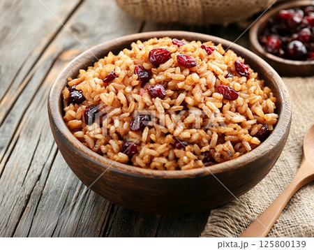 A wooden bowl filled with rice and cranberries on a rustic wooden table with a spoon beside it 125800139