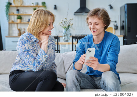 Middle aged mother and teenage son sitting together at home looking at smartphone Middle aged mother and teenage son sitting together at home looking at smartphone 125804027