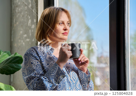 Portrait of beautiful middle-aged woman with cup in her hands looking out window 125804028