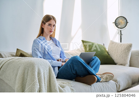 Portrait of young woman sitting on sofa with laptop looking at camera, in home interior 125804046