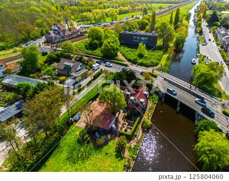 Aerial view of a railway crossing with cars and cyclists, a small station nearby, surrounded by lush green forest under a clear blue sky. Peaceful rural transportation scene. 125804060