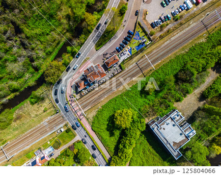 Aerial view of a railway crossing with cars and cyclists, a small station nearby, surrounded by lush green forest under a clear blue sky. Peaceful rural transportation scene. 125804066