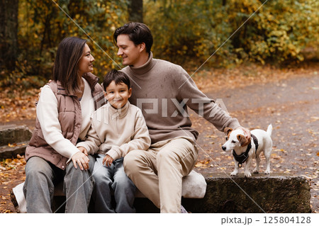 Happy family of three with dog sitting on bench in autumn park Happy family of three with dog sitting on bench in autumn park 125804128