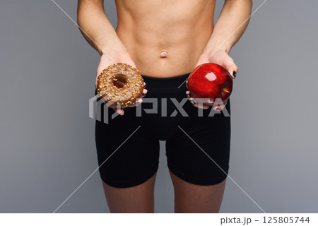 Sporty woman with a perfect body holding donut and apple. Healthy low calorie meals, fresh fruits Sporty woman with a perfect body holding donut and apple. Healthy low calorie meals, fresh fruits 125805744