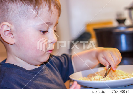 Small child with a spoon eating porridge from a large bowl in the kitchen in the evening 125808056