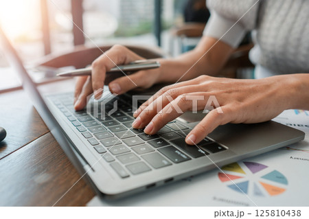 Close-up of hands typing on a laptop in a bright office setting with financial documents. 125810438
