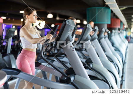 Young woman stands on simulator, performs fitness task using an elliptical simulator Young woman stands on simulator, performs fitness task using an elliptical simulator 125810443