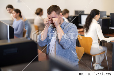 Male student rubs her tired eyes with her hands after working for long time at computer monitor in classroom Male student rubs her tired eyes with her hands after working for long time at computer monitor in classroom 125810669