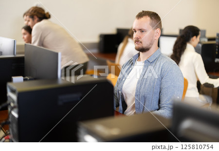 Guy student of computer courses looks at monitor screen, types on keyboard and does work Guy student of computer courses looks at monitor screen, types on keyboard and does work 125810754