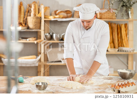 Guy works in bakery as baker, kneads dough, works with flour. 125810931