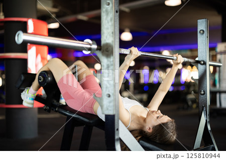 Girl lies on bench and lifts barbell, strengthens muscles of upper body 125810944
